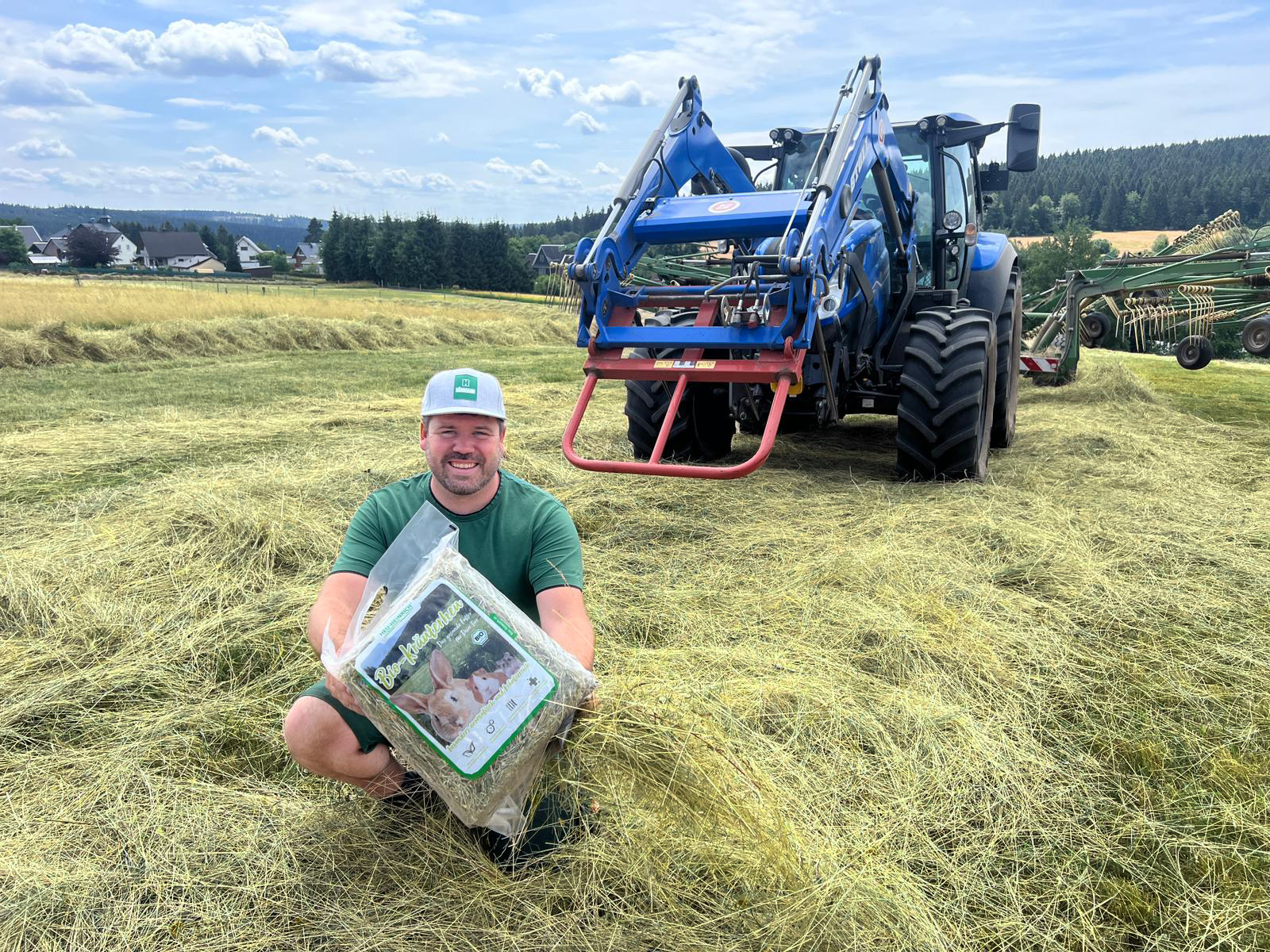 Landwirt mit Bio-Kräuterheu von Heu Heinrich auf gemähter Wiese vor Traktor – nachhaltige Heuernte in Thüringen