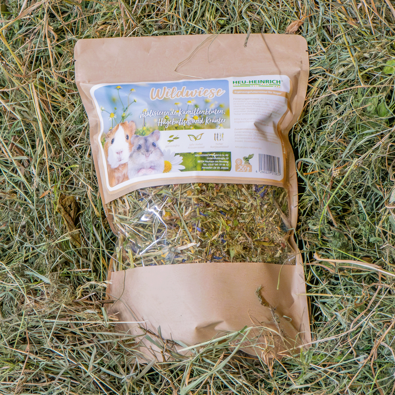 250g Futtermischung " Wildwiese " von Heu-Heinrich, Trockenfutter für Nager. Ergänzungsfuttermittel Foto von der braunen Papierverpackung mit Foliensichtfenster. Auf Heu liegend.
