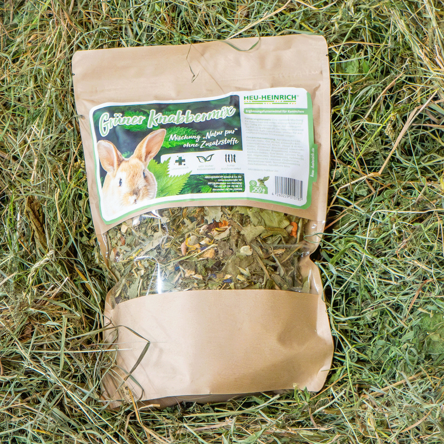 250g Futtermischung "Grüner Knabbermix" von Heu-Heinrich, Trockenfutter für Kaninchen. Ergänzungsfuttermittel Foto von der braunen Papierverpackung mit Foliensichtfenster. Auf Heu liegend.